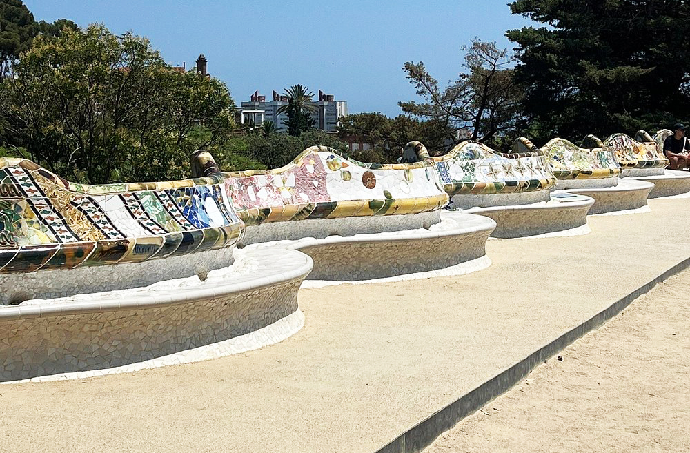 Os bancos da praça são revestidos com trencadís, formando desenhos coloridos e abstratos. Cada curva do banco foi pensada para o conforto dos visitantes. O mosaico reflete a luz do sol e cria um espetáculo visual único. Essa técnica reciclava pedaços de cerâmica quebrada, mostrando a visão sustentável de Gaudí. É um exemplo de arte aplicada ao cotidiano.