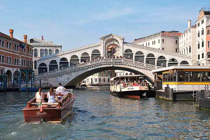 A Ponte de Rialto é a mais famosa de Veneza e uma das mais antigas, erguida em pedra no século 16 para substituir estruturas anteriores de madeira. Sua imponência reflete a prosperidade comercial da cidade, já que conecta o coração financeiro ao mercado central. O arco único, ladeado por lojas, tornou-se ícone arquitetônico e ponto de encontro para mercadores e viajantes. É um dos locais mais fotografados, reunindo turistas e moradores em sua travessia diária. A ponte simboliza a força econômica