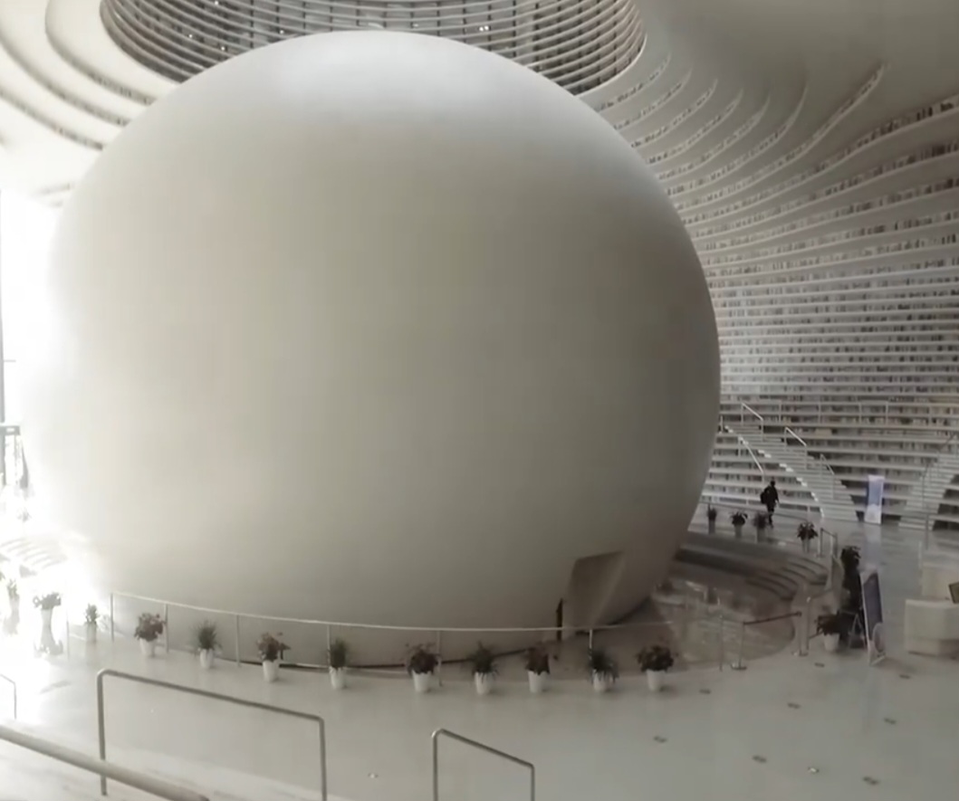 A luz natural invade o átrio e se reflete nas superfícies brancas, criando um ambiente quase futurista. No centro, o enorme auditório esférico, conhecido como “O Olho”, domina o espaço e é cercado pelas estantes onduladas que caracterizam a biblioteca.