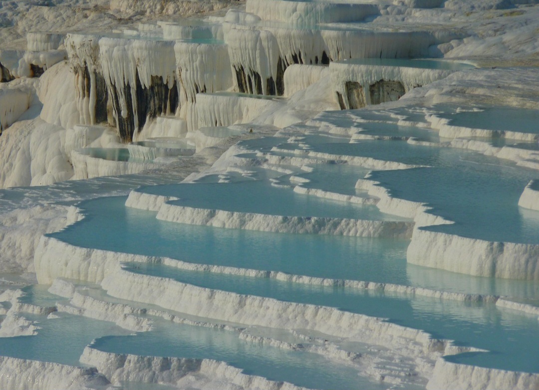 A água mineral percorre a encosta e deposita sucessivas camadas de calcário, criando pequenas barragens e piscinas em diferentes alturas. Esse processo geológico continua acontecendo, embora as formações levem muito tempo para se desenvolver. A combinação do branco intenso com os reflexos azulados da água torna a paisagem especialmente impressionante.